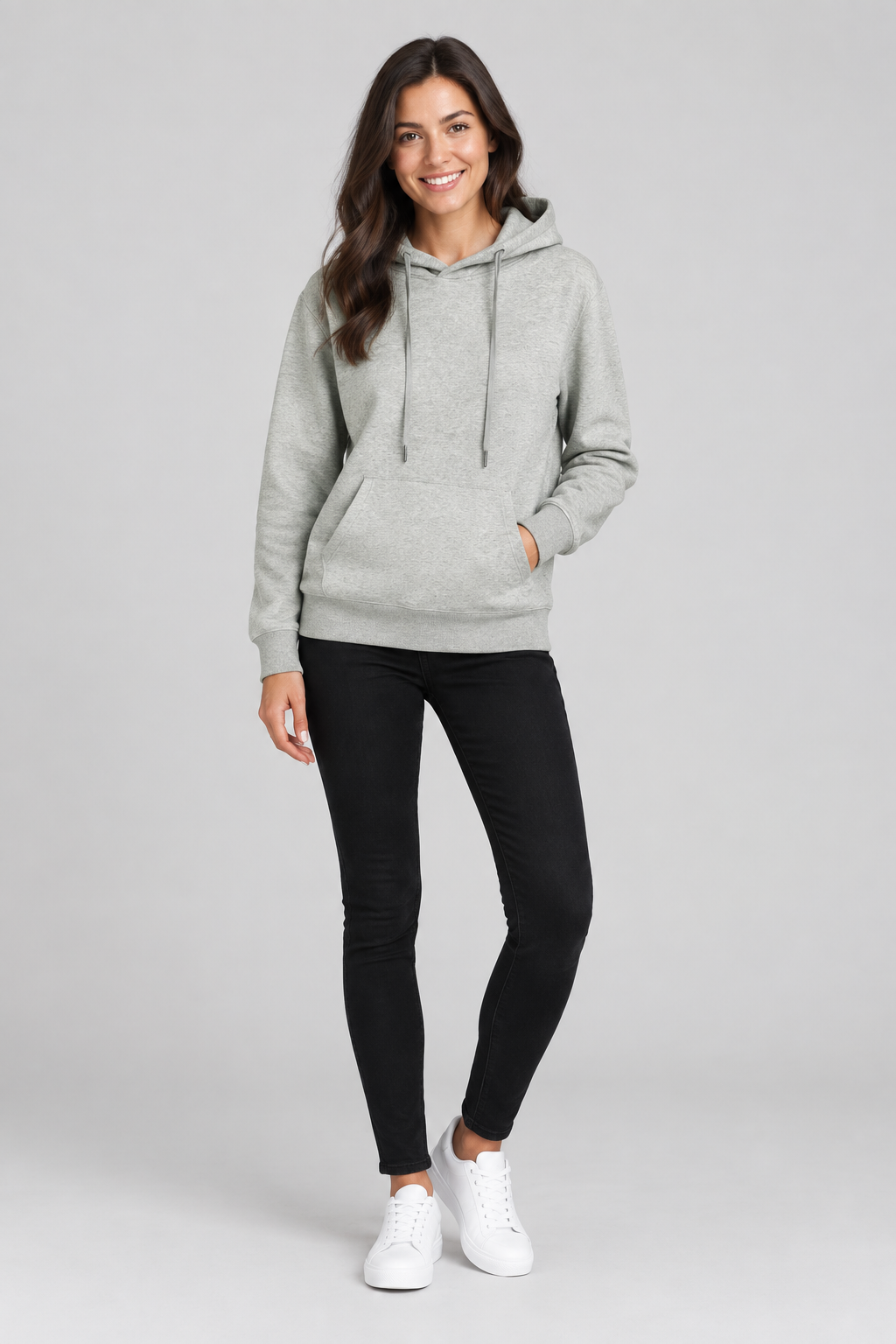 Full-body studio photo of a smiling brunette model wearing the hoodie, black jeans, and white shoes